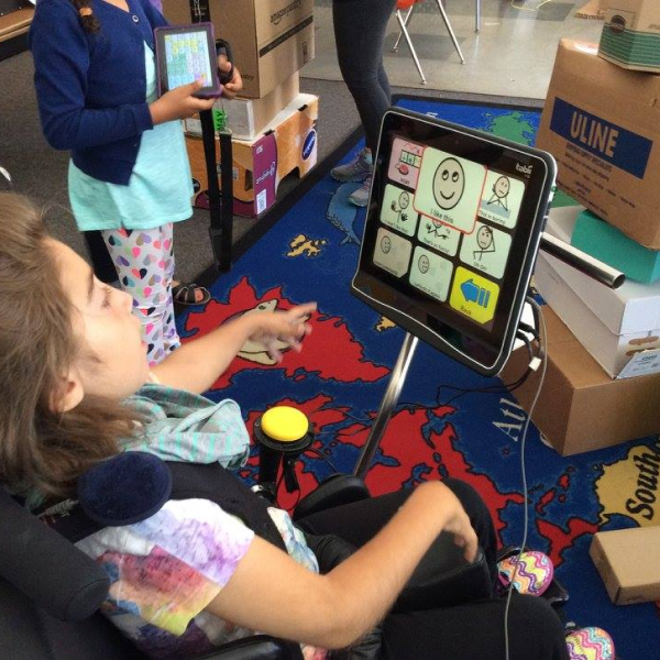 Image of a child in a wheelchair looking at an AAC device that has several images on it. An image of a smiling face is highlighted